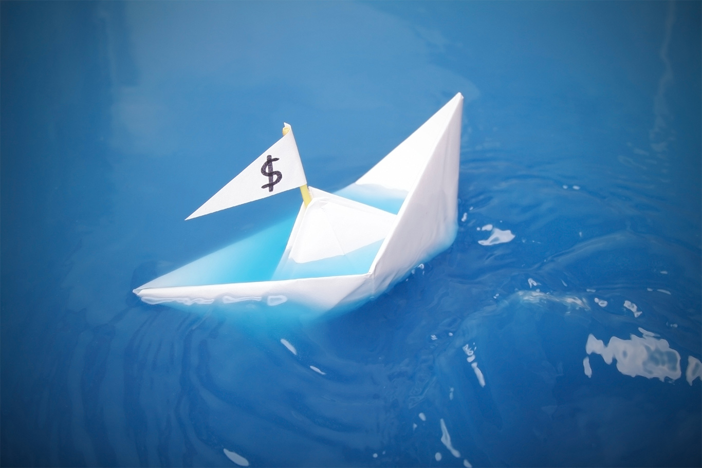 paper boat with a dollar sign on its flag is sinking in blue water